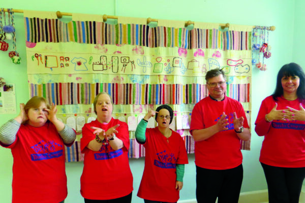 photo: 5 people are standing in a line in front of a wall hanging. They are all wearing matching red t-shirts with the DIY Theatre logo on. Each is doing a different sign with their hands.