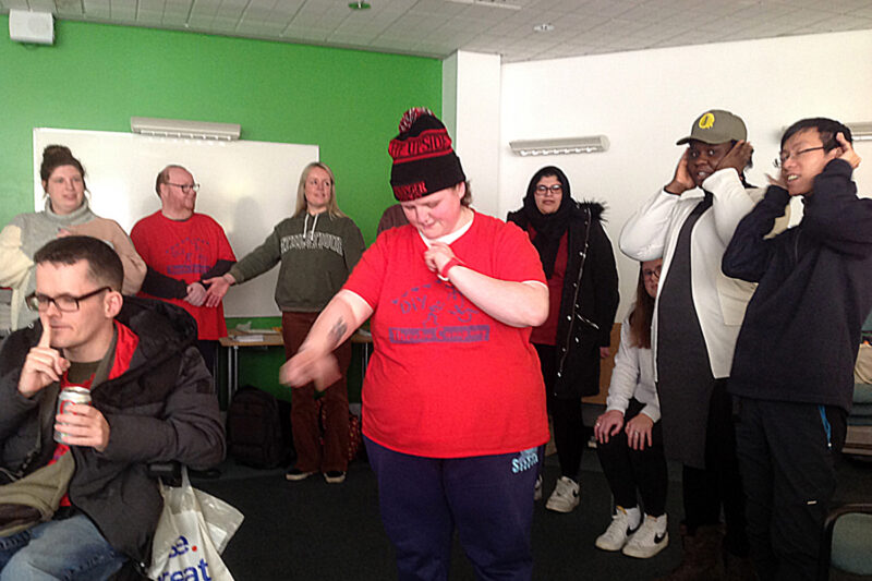 We can see a room full of people doing strange actions! A woman in the middle, wearing a DIY t-shirt is looking down whilst pointing up with her left hand and moving her right hand so quickly it's a blur. At the back we can see one person with their hands over their ears whilst another has their hands cupped behind their ears. A person sitting in a wheelchair at the front is holding a finger infront of their mouth 'shh'. Some other people are looking on.