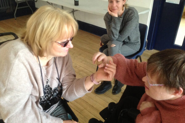 photo: there are two women close to each other, they are holding out and just touching each others hands tenderly. There is another woman looking on from further away, she has a gentle smile on her face.