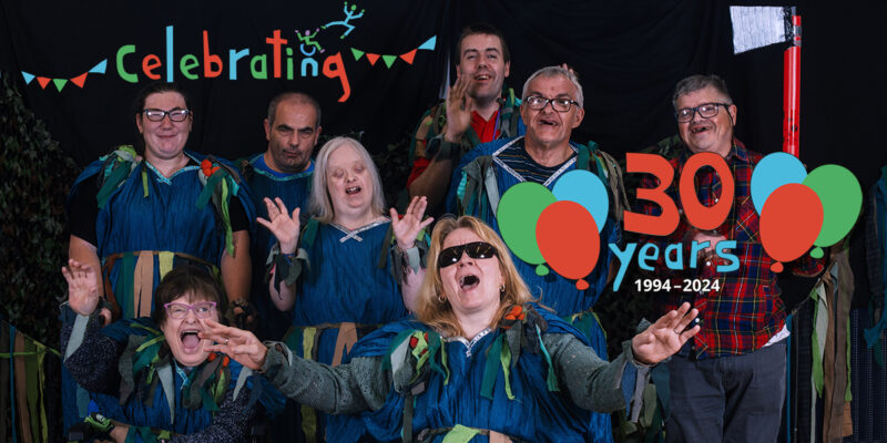 photo: there are a group of people in costumes. Most have blue tunics but one is wearing a red and blue workwear shirt. They are all facing the camera, many have their hands raised and are shouting or singing. At front it says 'Celebrating 30 years 1994-2024' in colourful letters with balloons