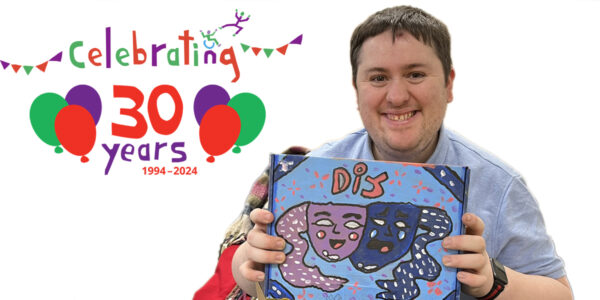 photo of a smiling young man holding the lid of a box open so that we can see it. The box is colourfully painted. In red it has "DIY" letters above sad and happy masks - the symbol of theatre.