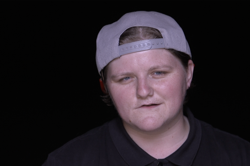 close up photo of a young woman. She is wearing a grey baseball cap back to front and is staring at the camera. The background is black and so is her t-shirt which makes the picture very dramatic and serious.