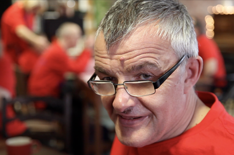 photo: close up of an older man with grey hair and glasses. He is wearing a bright red t-shirt as are others we can see in the background. He is looking over his glasses at us.