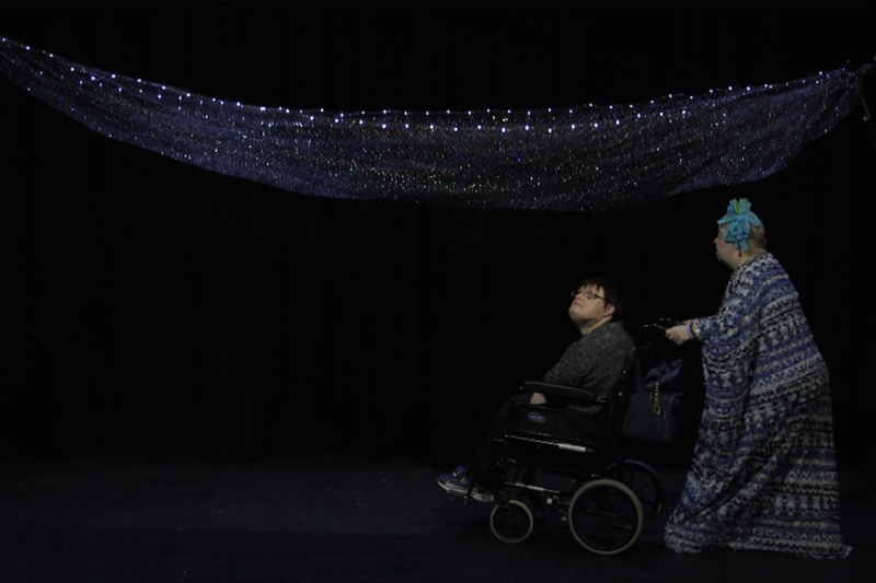 photo: we are looking at a dark scene, perhaps a stage. On the right a person dressed in a strange blue and white full length costume is pushing someone in a wheelchair to the left. Above them is a net of lights and the person sitting is looking up at these.