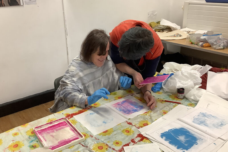 photo: A woman is sitting at a desk wearing blue rubber gloves and a shirt on backwards. The desk is covered in paper and it looks like she has been printing with blue and purple ink. Infront of her is a print of a face. Another woman is leaning towards her as she looks at the print.