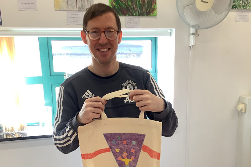 photo: A man is smiling and holding up a tote bag for us to see. The bag has a purple triangle on it. Inside this are lots of coloured starts and a figure with hands waving.