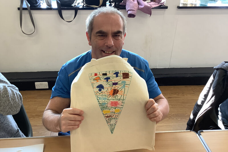 photo: A man is smiling and holding up a tote bag for us to see. The bag has a triangle on it made up of lots of colourful drawings.