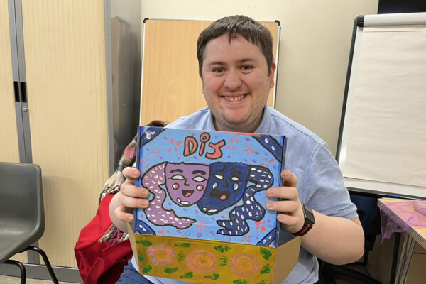 photo: a young man is smiling at us. In his hands he has a box which he is holding open. we can't see what is in the box but we can see the lid. This is covered in a painting of the masks that represent theatre and "DIY" in red letters.