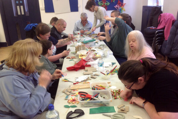 photo: people are sitting around a long table. The table is covered in coloured paper, scissors, pens and the people are making things with these.