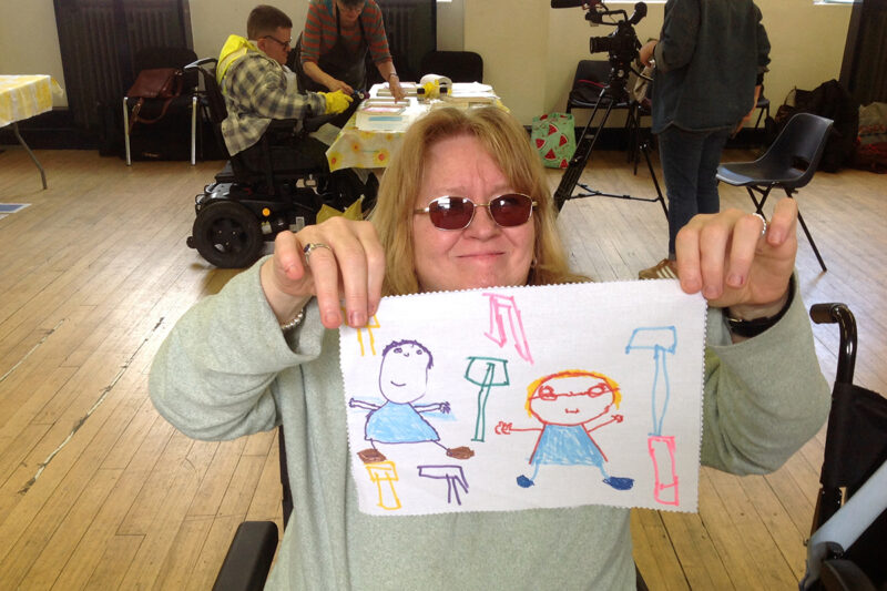 photo: a smiling woman is holding up a piece of white fabric with a brightly coloured drawing on it of people and tall objects. Behind her other people are working at a table and filming.