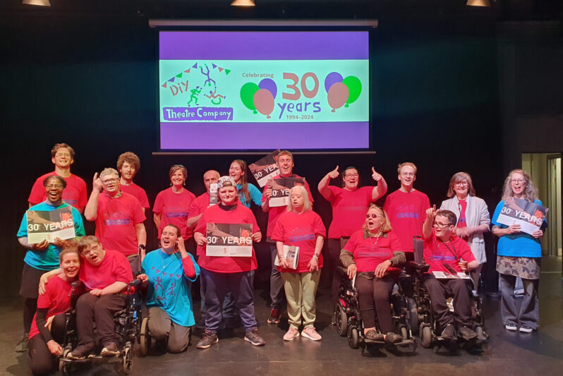 photo - lots of people are on a stage. Most are wearing red t-shirts. Everyone looks happy and many are holding brochures which say '30 YEARS' Behind them there is a large screen which has DIY's Logo and '30 years' with balloons around.