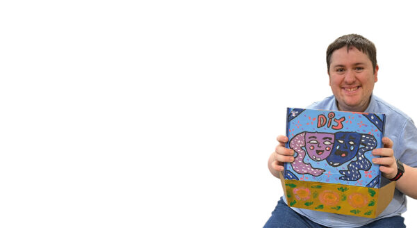 photo of a smiling young man holding the lid of a box open so that we can see it. The box is colourfully painted. In red it has "DIY" letters above sad and happy masks - the symbol of theatre.