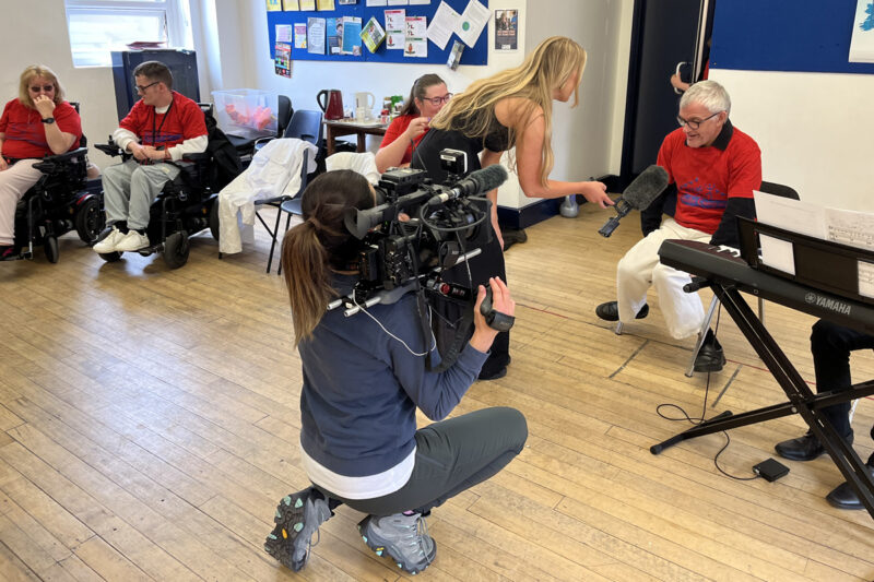 photo: people in red t-shirts are sitting along the edge of a room. In the foreground a young person is kneeling down and holding a big camera on their shoulder. They are filming. A young woman with long blond hair is holding a mike towards an older man in glasses.