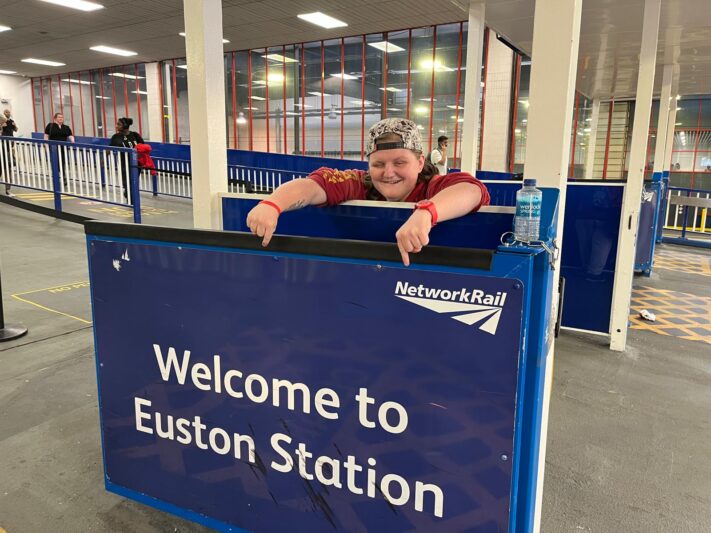 charlotte is smiling and pointing down to a sign that says welcome to euston station she is inside the train station in london