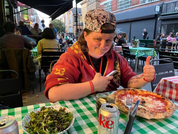 charlotte is sitting at a table which has pizza and lots of tasty food on it she is smiling with her right hand thumb up showing she is happy and her left hand is holding her phone she has a grey hat on backwards and i red hoody on