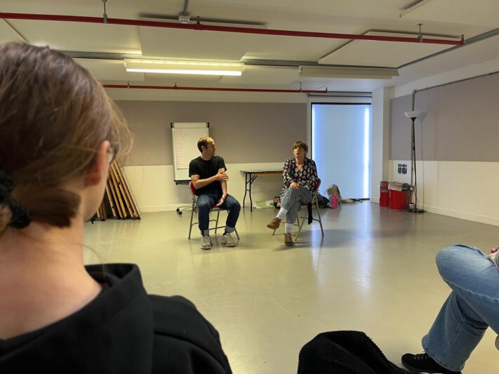 two people are sat on chairs talking and they look like they are in the middle of acting we can see the side of someones head closer to the camera who is watching the two who are talking 