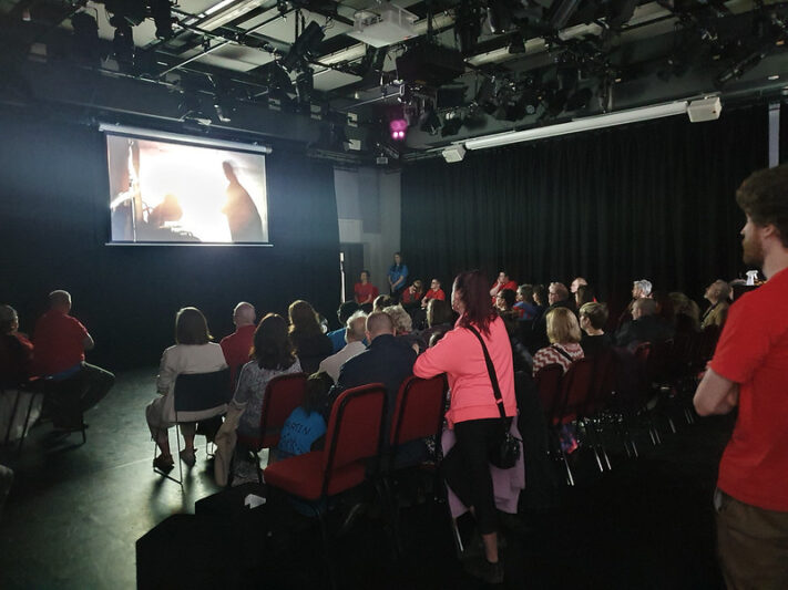 A seated audience watches a projected video in a dimly lit studio space with black curtains and stage lighting. The scene on the screen features silhouetted figures, creating a dramatic visual effect.