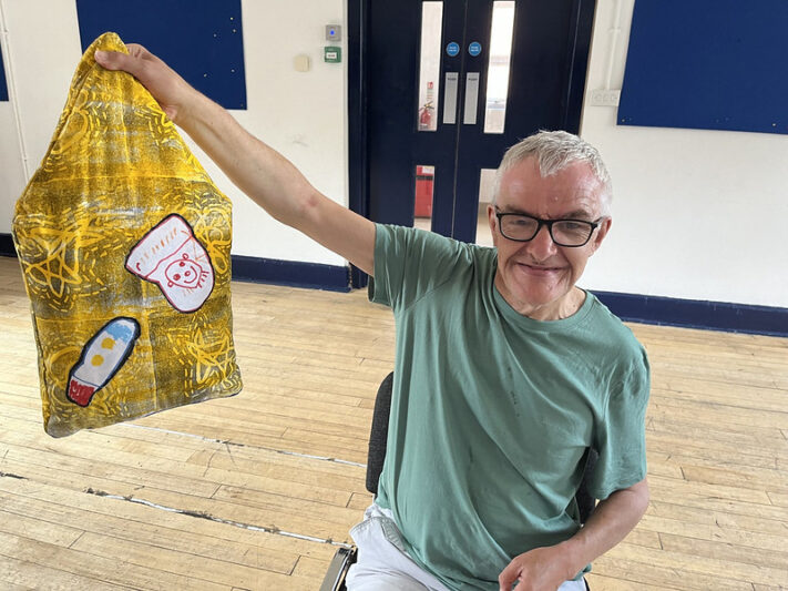 A smiling individual holds up a vibrant, hand-decorated yellow bag featuring artistic designs, including a drawing of a cat and a colorful rocket.