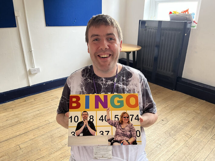 A smiling individual holds up a colorful bingo card featuring vibrant letters spelling "BINGO" and images of two people gesturing enthusiastically.