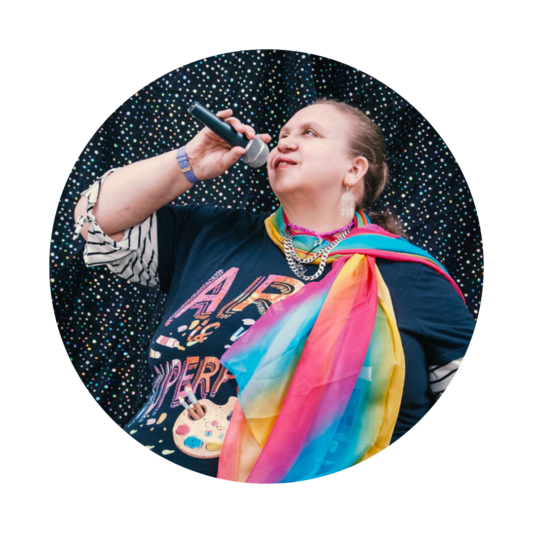 A woman sitting on a chair, holding a microphone to her mouth as she looks upward with a smile. She is wearing a colorful outfit, including a black T-shirt with the text 'Art and Expression,' a striped long-sleeve top underneath, and a vibrant rainbow scarf draped around her shoulders. A decorative banner with a treble clef and floral design hangs from her lap. The background features a sparkly black curtain, adding a celebratory atmosphere.