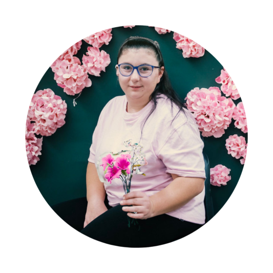 A woman sitting in front of a green backdrop decorated with clusters of pink flowers. She is wearing glasses, a pale pink T-shirt, and black trousers, holding a small bouquet of pink flowers. She looks directly at the camera with a calm expression.