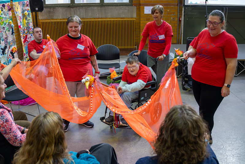 A group of individuals in red T-shirts engage in a lively activity with an orange fabric stretched between them, creating a dynamic and inclusive atmosphere.