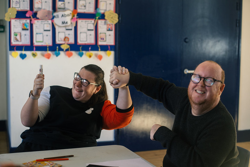 Two individuals sit at a table smiling warmly, one holding a marker and the other raising their hand in a playful gesture of celebration or victory.