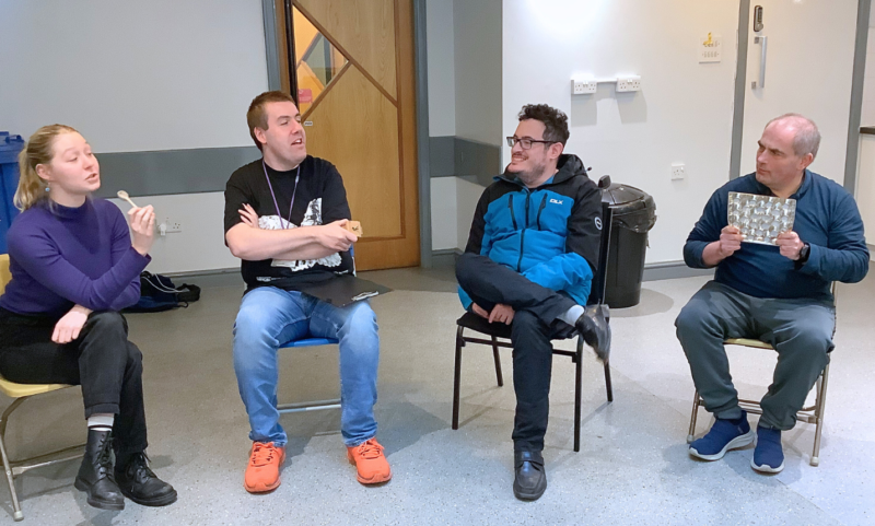 Four people sitting in a semi-circle on chairs in a brightly lit room. The person on the far left is holding a small object, possibly a spoon, and appears to be speaking. The second person, wearing an illustrated black T-shirt and orange trainers, gestures with their hand while speaking. The third person, in a blue jacket and glasses, sits cross-legged and smiles. The person on the far right, in a blue long-sleeve top, holds up a silver reflective sheet. The background features a wooden door, a recycling bin, and a kitchen area.