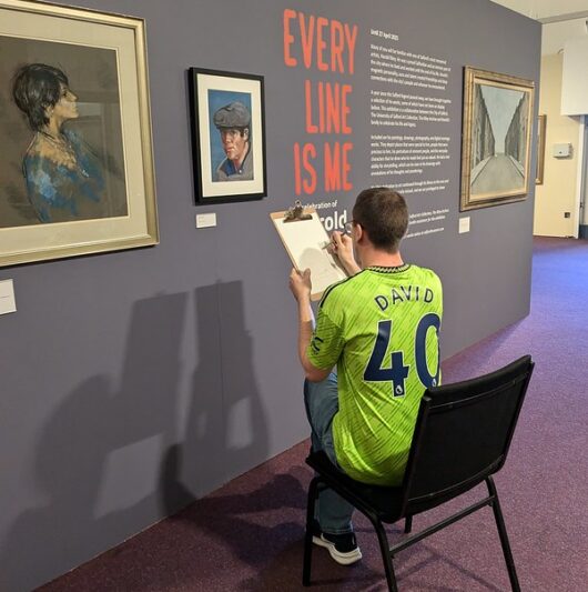 David, wearing a bright green football jersey with his name and the number 40 on the back, sits on a chair in a museum gallery sketching on a clipboard. He is positioned in front of a wall display titled 'EVERY LINE IS ME,' surrounded by framed artwork. The atmosphere is calm and focused, emphasising creativity and engagement with the artwork.