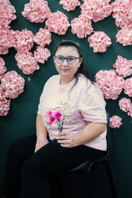 A woman with long dark hair tied back, wearing blue glasses, a light pink top, and black trousers, sits on a black chair. She is holding a small bouquet of pink and white flowers and is surrounded by a vibrant backdrop of pink hydrangea-style flowers on a dark green background.