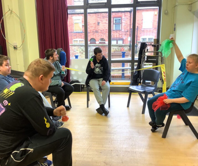 A group of five participants sit in a semi-circle in a bright room with large windows. Each person holds a colourful prop, such as maracas or scarves, engaging in an interactive activity. The participants appear focused and collaborative, while the room’s natural light highlights the vibrant energy of the session.