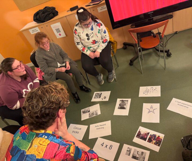 Four participants sit in a semi-circle, engaged in a group activity. They are focused on printed images and phrases laid out on the floor, such as 'Circus tricks,' 'Arts Award training,' 'Lived experience leadership,' and a large star illustration. A facilitator with curly hair, wearing a colourful patterned shirt, sits with their back to the camera, observing the discussion. The setting is a bright room with orange walls, a large screen, and minimal furniture, creating a collaborative and creative atmosphere.